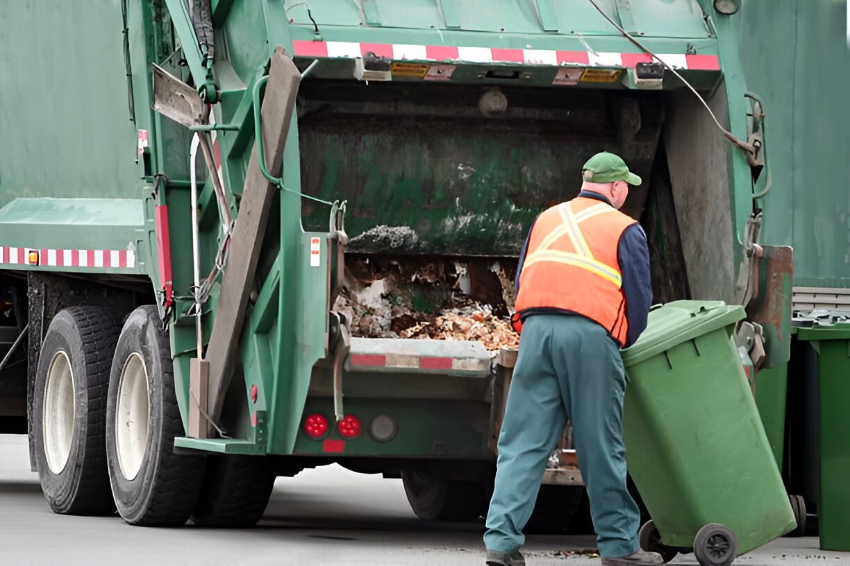 Waste management worker positions green bin on the automatic dumper at the back of truck.
